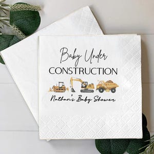 Op de afbeelding: Witte papieren servetten met een babyshower thema. De servetten tonen de tekst "Baby Under Construction" met illustraties van bouwmachines. De tekst "Nathan's Baby Shower" is ook opgenomen.