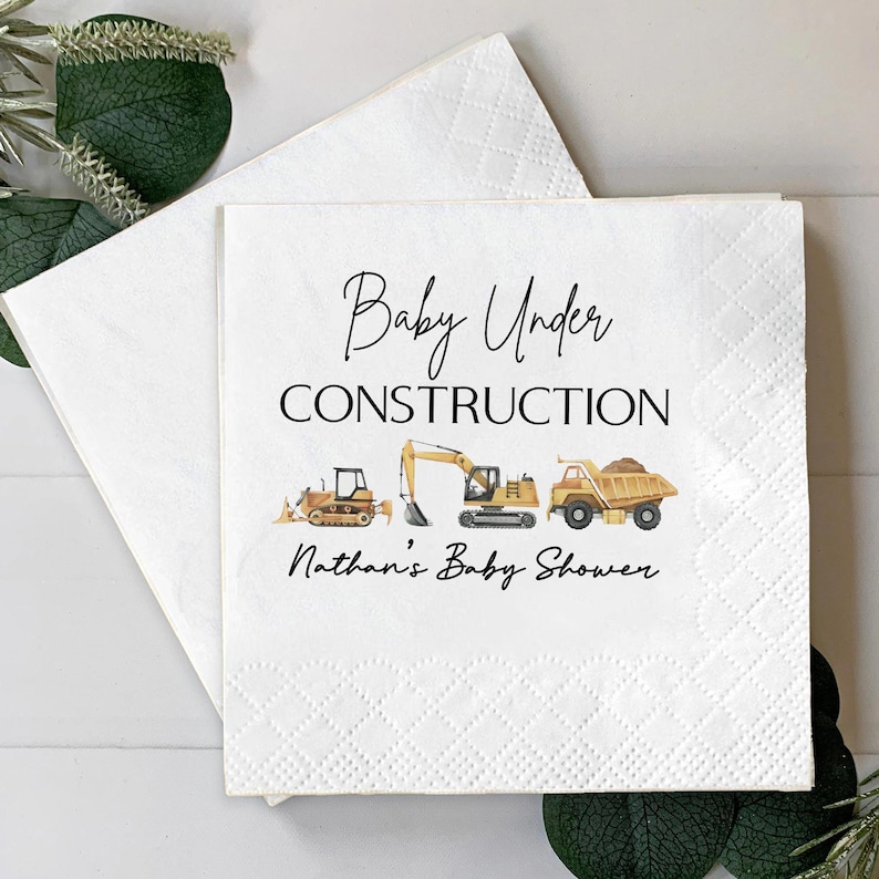 Op de afbeelding: Witte papieren servetten met een babyshower thema. De servetten tonen de tekst "Baby Under Construction" met illustraties van bouwmachines. De tekst "Nathan's Baby Shower" is ook opgenomen.