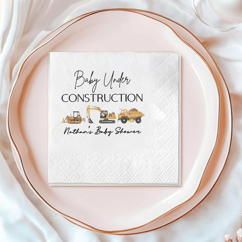 Op de afbeelding: Witte vierkante papieren servet met de tekst "Baby Under Construction" en "Nathan's Baby Shower". De servet toont illustraties van gele en bruine bouwmachines, waaronder een dumper, graafmachine en wiellader.