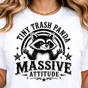 May include: White t-shirt featuring a black and white raccoon illustration with the text "Tiny Trash Panda Massive Attitude". The raccoon is centered with its arms raised, surrounded by stars and a rainbow arc.