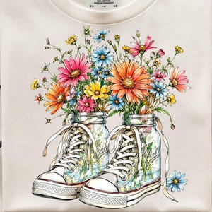 May include: A cream-coloured t-shirt with a colourful floral design. The graphic shows a pair of trainers filled with bright flowers. The shirt has a crew neck and short sleeves. The tag reads "COMFORT COLORS".