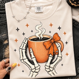 May include: A cream-colored t-shirt featuring a graphic of two skeletal hands holding an orange mug with a bow. The mug contains a dark liquid with rising steam. The design is surrounded by small black and orange four-pointed star icons.