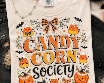 以下が含まれることがあります： オレンジと黒で「CANDY CORN SOCIETY」という文字がプリントされたクリーム色のTシャツ。デザインには、笑顔のキャンディコーンのキャラクター、オレンジ色のカボチャ、チェック柄のリボン、小さな黒いコウモリのイラストが描かれています。