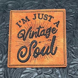 May include: A square-shaped orange patch with a textured surface and a stitched border. The patch features black embroidered text that reads I'M JUST A VINTAGE SOUL, accompanied by a small arrow graphic and radiating lines centered on the word Vintage.