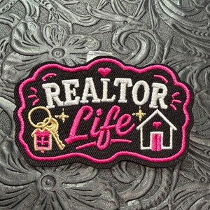 May include: A black embroidered patch with a scalloped pink border featuring the white text REALTOR and the pink script text Life. The design includes a gold key with a pink house keychain and a white house icon with a pink heart and door.
