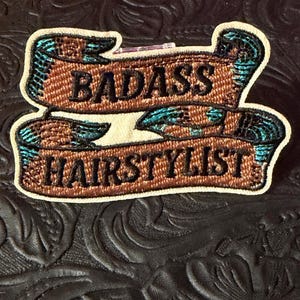 May include: An embroidered patch featuring two brown ribbon-style banners with black stitched text that reads BADASS HAIRSTYLIST. The design includes teal and black accents on the edges of the ribbons and a cream-colored border surrounding the entire shape.