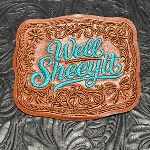 May include: A rectangular brown leather patch with rounded corners featuring intricate copper-colored embroidery of floral scrolls and bows. The center displays the text Well SHEEYit embroidered in a bright turquoise blue script font.