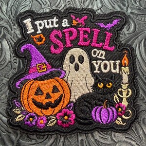 May include: A black embroidered patch featuring the text I put a SPELL on YOU in pink and white thread. The design includes a jack-o'-lantern wearing a purple witch hat, a white ghost, a black cat, a lit candle, and small purple flowers.