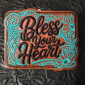 May include: A rectangular brown leather patch featuring black embroidered text that reads Bless your Heart. The patch is decorated with intricate turquoise floral and scrollwork embroidery along the edges, framed by a stitched border.