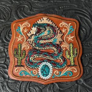 May include: A tan leather patch featuring an embroidered design of a coiled snake in shades of teal and maroon. The patch includes two green cacti, tan stars, and decorative scrollwork, centered with a teal oval stone surrounded by white beads.