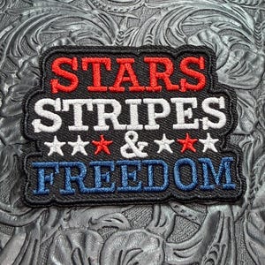 May include: A rectangular embroidered patch with a black background featuring the red text STARS, white text STRIPES, a white ampersand flanked by two white stars and two red stars, and blue text FREEDOM, all bordered by a thick black stitched edge.