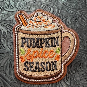 May include: An embroidered patch shaped like a mug filled with a beverage topped with whipped cream and a cinnamon stick. The mug features the text PUMPKIN spice SEASON in black and orange thread, accented with small green and orange leaf icons.