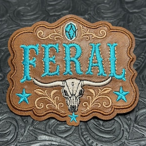 May include: A brown leather patch with scalloped edges featuring the word FERAL embroidered in bright turquoise thread. The design includes a white steer skull, a small turquoise oval accent, and three turquoise stars with tan scrollwork.
