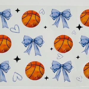 May include: Sheet of stickers featuring orange basketballs, light blue bows, black star shapes, and white heart shapes. The basketballs have black lines. The bows have a watercolor effect. The stickers are on a clear background.