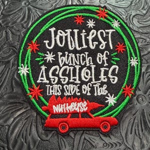 May include: A circular embroidered patch with a black background featuring a green wreath, red and white star accents, and the text "JOLLIEST bunch of ASSHOLES THIS SIDE OF THE NUTHOUSE" above a red station wagon carrying a Christmas tree.