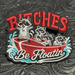 May include: An embroidered patch featuring three raccoons in a red boat on blue water. The text BITCHES is stitched in red at the top, and the text Be Floatin is stitched in white at the bottom. The patch has a black background and border.