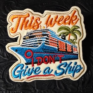 May include: An embroidered patch featuring a blue cruise ship, a green palm tree, and the text "This week I DON'T Give a Ship" in orange and blue thread. The design is set against a cream-colored background with a die-cut border shape.