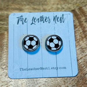 May include: A pair of soccer ball stud earrings with a black and white design. The earrings are on a light blue card with the text "The Leather Nest" and "TheLeatherNest1.etsy.com". The background is a wooden surface.