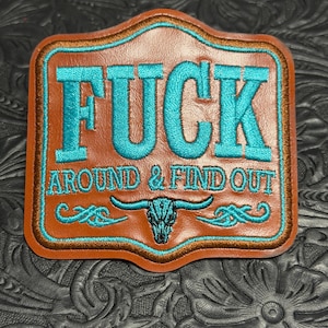 May include: A brown leather patch with a scalloped edge featuring the embroidered turquoise text FUCK AROUND & FIND OUT. Below the text is a turquoise embroidered steer skull graphic with decorative scrollwork on either side of the skull.