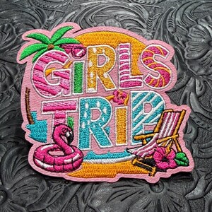May include: An embroidered patch featuring the text GIRLS TRIP in colorful, patterned letters. The design includes a green palm tree, a pink flamingo pool float, a yellow sun, a beach chair, and a pink hibiscus flower on a light pink background.