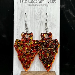 May include: A pair of arrowhead-shaped earrings with red, orange, and gold glitter embedded in clear resin. The earrings are on a white wooden display stand with the text "The Leather Nest Handmade Jewelry".