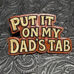 May include: A brown leather patch with embroidered gold lettering that reads "PUT IT ON MY DAD'S TAB". The patch features a thick black border around the text and the leather edge, with the letters filled in gold thread and outlined in black.