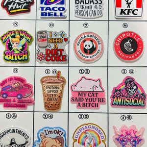 May include: A collection of colorful, glittery acrylic charms featuring various designs. Includes fast food logos like Pizza Hut and KFC, along with humorous phrases and images such as "My Cat Said You're A Bitch" and a cartoon fairy.