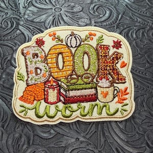 May include: An embroidered patch featuring the text BOOK WORM in colorful, patterned letters. The design includes a stack of books, a pair of glasses, a lit candle, a mug with a heart, and a pumpkin, all accented with autumn-themed foliage.