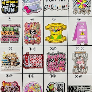 May include: A collection of decorative charms with various designs and text. The charms feature phrases like "When God Made Me He Grinned," "How You Doin?" and "I'm Made of Glitter Water and Anxiety." Colors include black, white, pink, and green.