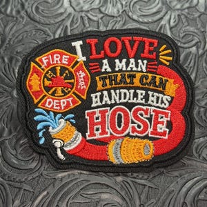 May include: A black embroidered patch featuring a red and gold fire department emblem, a red fire hose spraying blue water, and the text I LOVE A MAN THAT CAN HANDLE HIS HOSE in red, white, and yellow thread on a black background.