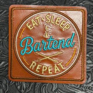 May include: A square brown leather patch featuring circular embroidery. The text EAT SLEEP REPEAT is stitched in gold, with the word Bartend in bright blue script. A gold jigger and crossed bar spoons are embroidered in the center of the design.
