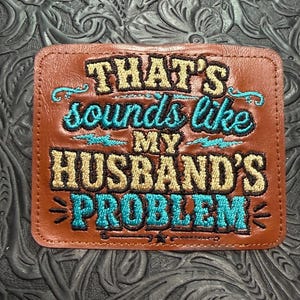 May include: A rectangular brown leather patch featuring embroidered text that reads THAT'S sounds like MY HUSBAND'S PROBLEM. The words are stitched in a combination of gold and bright turquoise thread with decorative flourishes and lines.