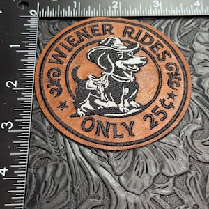 May include: A circular brown leather patch featuring a black embroidered graphic of a dachshund wearing a cowboy hat and saddle. The text reads "WIENER RIDES" and "ONLY 25¢". A black metal ruler is visible along the top and left edges for scale.
