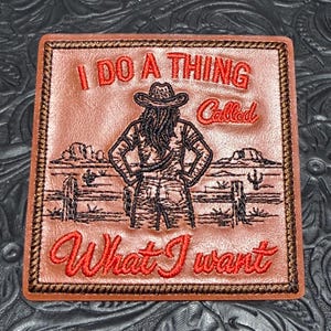 May include: A square brown leather patch featuring dark brown embroidered stitching around the border. The center displays a desert landscape with a person wearing a cowboy hat, with red embroidered text reading I DO A THING Called What I want.