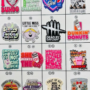 May include: A collection of colorful, glittery charms with various designs and text. The charms include phrases like "I love Bunco," "Pepsi Girl," and "Dunkin' Donuts." Other charms feature cartoon characters and humorous sayings.