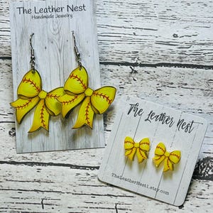 May include: Softball-themed earrings and stud earrings. The earrings are yellow with red stitching, shaped like bows. The stud earrings are smaller versions of the same design. The cards read "The Leather Nest Handmade Jewelry" and "TheLeatherNest1.etsy.com".