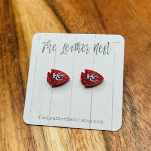 May include: A pair of red glitter earrings with a white "KC" logo on each earring. The earrings are on a white card with the text "The Leather Nest" and "TheLeatherNest1.etsy.com".