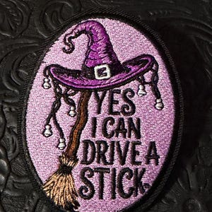 May include: An oval embroidered patch with a light purple background featuring a purple witch hat and a brown broomstick. The text reads YES I CAN DRIVE A STICK in black embroidered letters. The patch has a black border and small white bells.