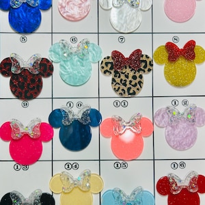 May include: A grid of sixteen Minnie Mouse-shaped acrylic charms in various colors and patterns. Each charm features a glittery bow and a different base color, including blue, pink, red, and yellow. The charms are arranged on a white grid.