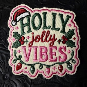 May include: An embroidered patch with a cream background featuring the text HOLLY in dark green, jolly in red, and VIBES in pink. The design includes a red Santa hat, green holly leaves with red berries, and a string of red, green, and pink lights.