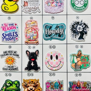 May include: A collection of colorful acrylic charms with various designs. Includes a yellow duck with sunglasses, a paw print, a smiley face, and text like "Howdy" and "Pray Through The Chaos".