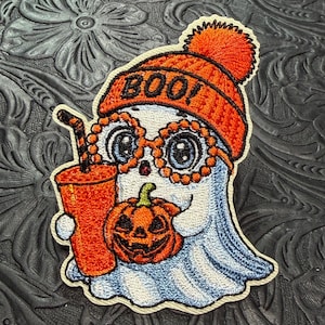 May include: An embroidered patch featuring a white ghost wearing an orange knit beanie with a pom-pom and the text BOO! The ghost wears orange circular glasses, holds an orange cup with a straw, and carries a small orange jack-o'-lantern pumpkin.