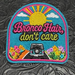 May include: An embroidered patch with a turquoise background featuring a yellow sun, white clouds, and a black SUV driving on a road. The text reads "Bronco Hair, don't care" in pink and yellow. Colorful flowers are on both sides of the road.