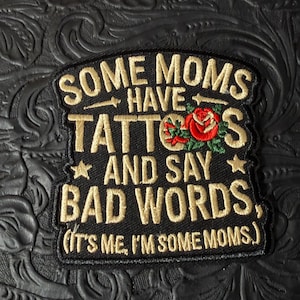 May include: A black embroidered patch featuring gold text that reads SOME MOMS HAVE TATTOOS AND SAY BAD WORDS, (IT'S ME, I'M SOME MOMS.). The design includes a red rose with green leaves and two small gold stars flanking the central text.
