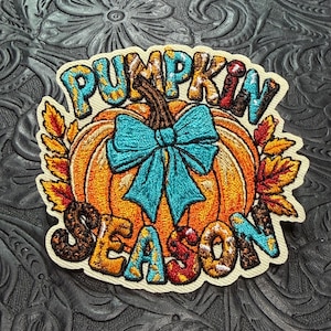 May include: An embroidered patch featuring a central orange pumpkin with a brown stem and a bright turquoise bow. The text PUMPKIN SEASON is stitched in multi-colored, patterned lettering above and below the pumpkin, flanked by autumn leaves.