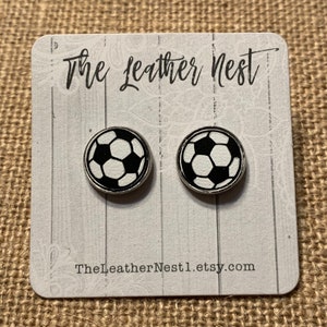 May include: A pair of black and white soccer ball earrings on a white card with the text "The Leather Nest" and "TheLeatherNest1.etsy.com".
