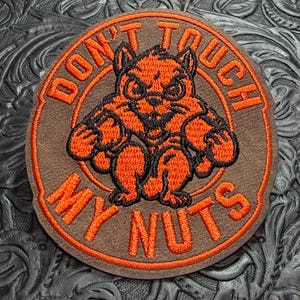 May include: A circular embroidered patch featuring a brown squirrel with an aggressive expression, holding two nuts. The text DON'T TOUCH MY NUTS is stitched in bright orange thread around the squirrel, set against a brown felt background.