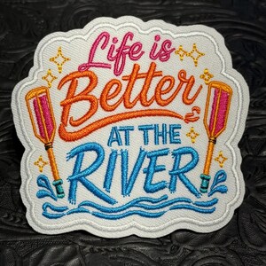 May include: A white embroidered patch with a scalloped edge featuring the text Life is Better AT THE RIVER in pink, orange, and blue thread. The design includes two orange and pink oars on the sides and small yellow star accents throughout.