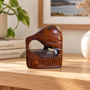 May include: A dark brown, polished wooden sculpture of a kiwi bird in a stylized, abstract form. The carving features smooth, rounded contours, a circular cutout representing the body, and linear etched details along the base and upper section.