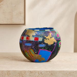 May include: A round, black vase featuring a vibrant, abstract design with gold leaf accents, bright blue rectangular shapes, red and white checkered patterns, and neon pink outlines. The surface displays a textured, layered artistic finish.
